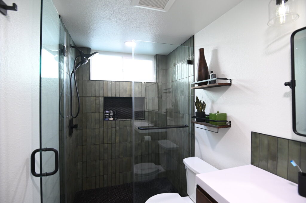 Modern bathroom with dark green vertical tile walk-in shower, black matte fixtures, and floating shelves by RnD Builders Inc