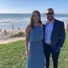 Couple in formal attire stands on a coastal overlook, overlooking a beach with people.