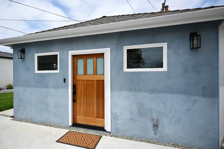 ADU Exterior with Blue-gray Stucco ADU exterior with blue-gray stucco, natural wood craftsman door, and white-trimmed windows by RnD Builders Inc