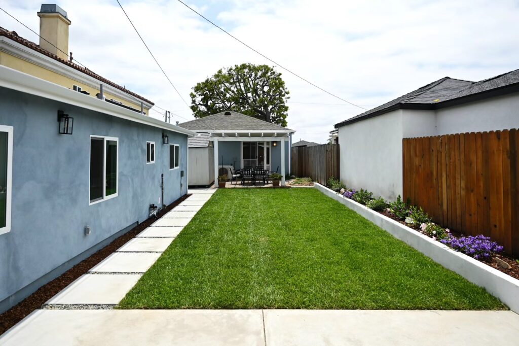 Landscaped Backyard with Green Lawn Landscaped backyard with green lawn, concrete stepping stones, white planter borders, and covered patio pergola by RnD Builders Inc