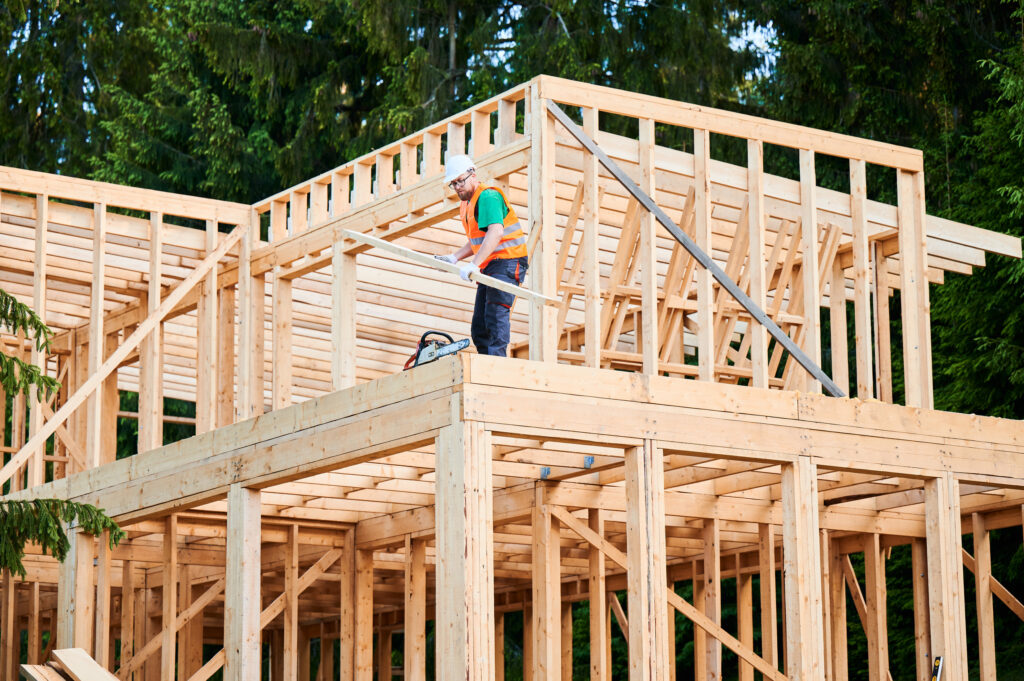 Carpenter holding large beam while builds wooden frame house near the forest.