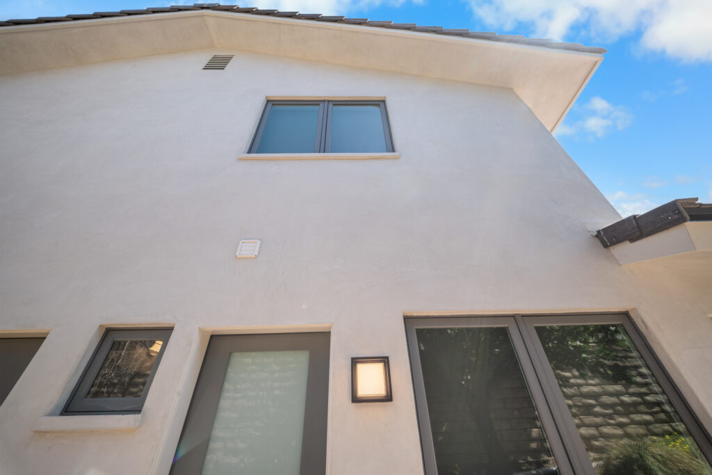 Two-story stucco home exterior with dark aluminum-framed windows and modern wall sconce by RnD Builders Inc