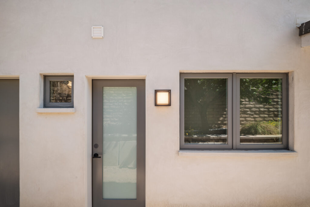 Modern stucco exterior with frosted glass entry door, dark gray window frames, and square wall sconce by RnD Builders Inc