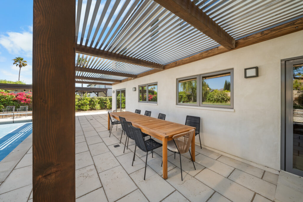 Covered outdoor patio with louvered pergola, teak dining table, and pool view by RnD Builders Inc