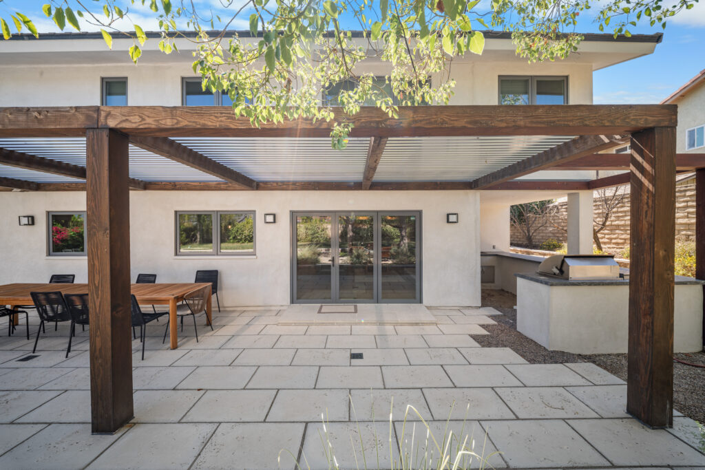 Modern covered patio with dark wood pergola, large format pavers, and outdoor kitchen by RnD Builders Inc