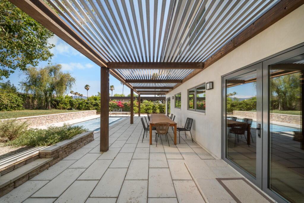Covered patio with louvered pergola, outdoor dining set, and poolside stone retaining wall by RnD Builders Inc