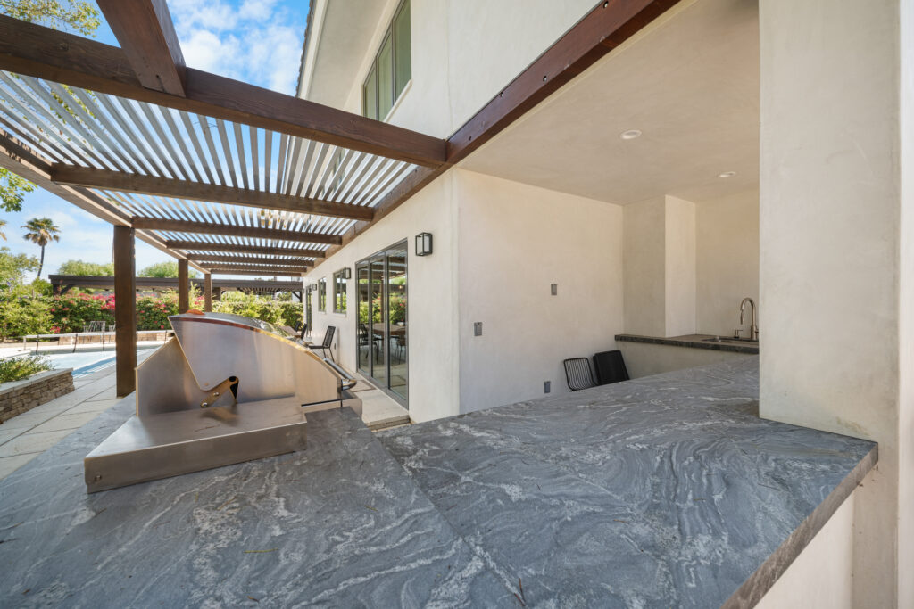 Covered outdoor kitchen patio with stone countertop, wood pergola, and pool view by RnD Builders Inc