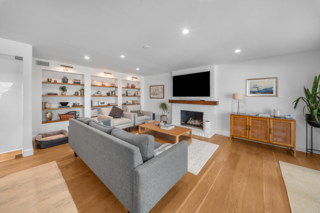 Bright living room with white brick fireplace, built-in shelves, and wide-plank hardwood floors by RnD Builders Inc