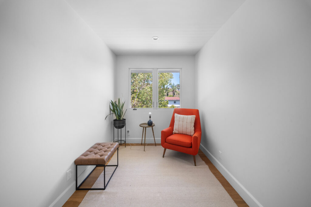 Minimalist sitting room with orange mid-century accent chair, tufted leather bench, and hardwood floors by RnD Builders Inc