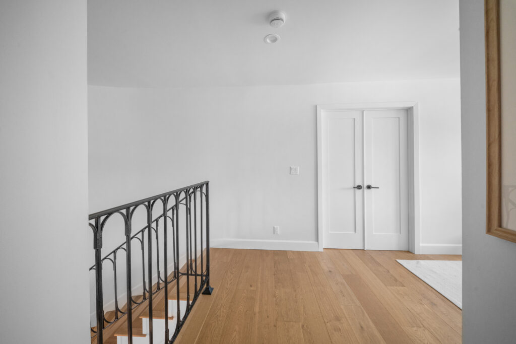 Upper hallway landing with wrought iron stair railing, white double doors, and natural oak hardwood flooring by RnD Builders Inc