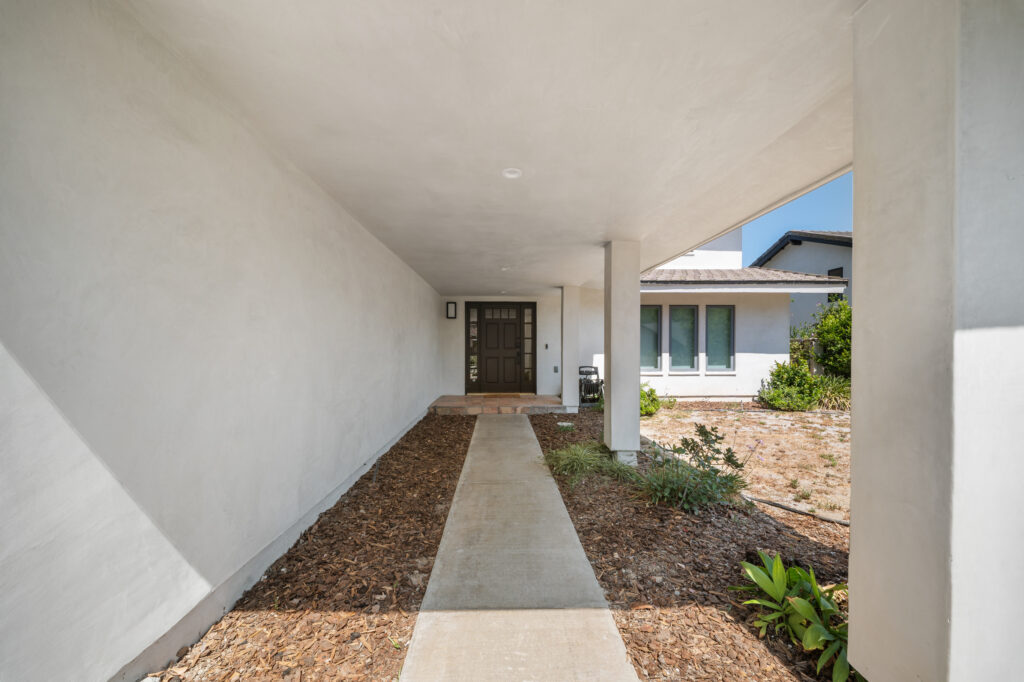 Covered front entry walkway with concrete path, wood mulch landscaping, and dark wood door by RnD Builders Inc