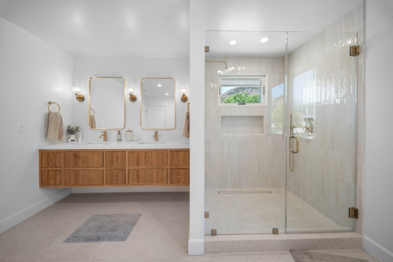 Bright primary bathroom with floating fluted wood vanity, dual gold mirrors, and frameless glass shower by RnD Builders Inc