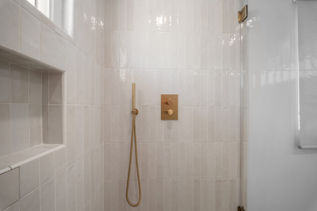 Walk-in shower with vertical white glossy tile, brushed brass handheld shower and thermostatic valve by RnD Builders Inc