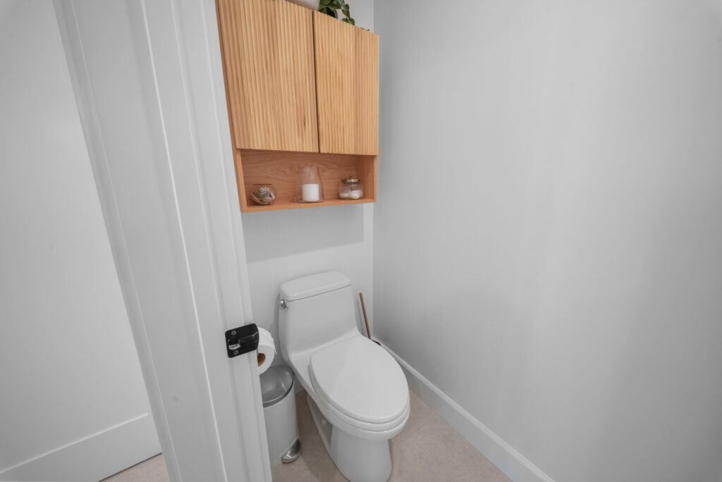 Compact bathroom water closet with white toilet and fluted wood upper cabinet by RnD Builders Inc