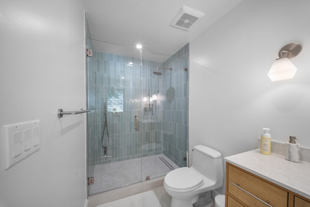 Modern bathroom with frameless glass shower, blue-green vertical subway tile, and warm wood vanity by RnD Builders Inc