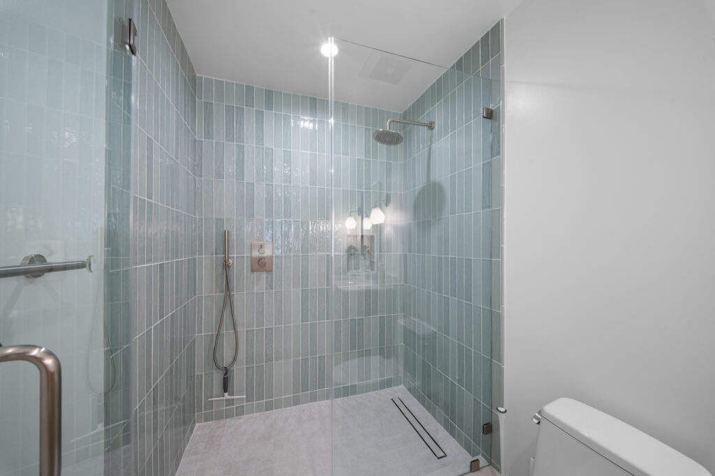 Walk-in shower with vertical sage green tile, frameless glass enclosure, and rain showerhead by RnD Builders Inc