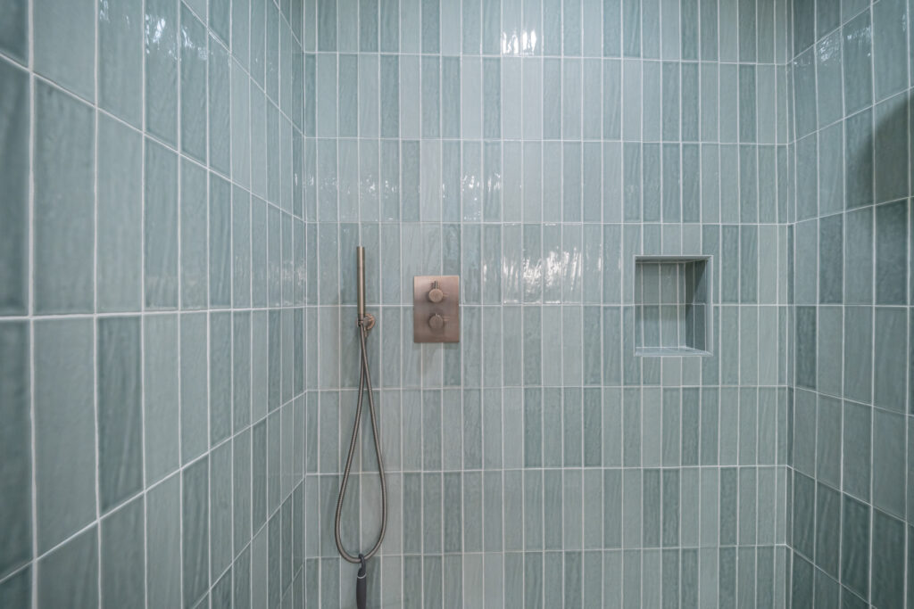 Walk-in shower with vertical sage green glossy tile, brushed bronze thermostatic valve, handheld wand, and recessed niche by RnD Builders Inc