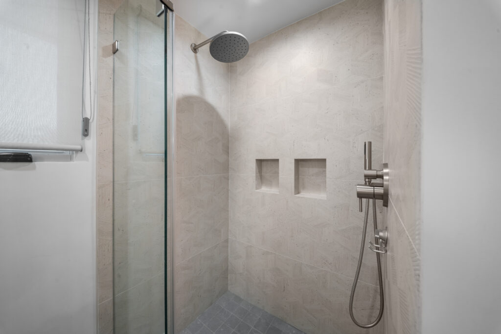 Walk-in shower with textured beige large-format tile, rain showerhead, and dual built-in niches by RnD Builders Inc
