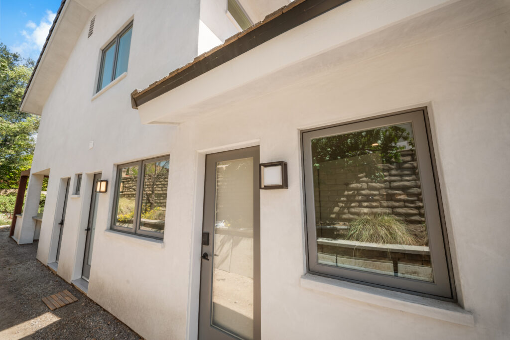Two-story white stucco home exterior with gray-framed windows and glass entry door by RnD Builders Inc