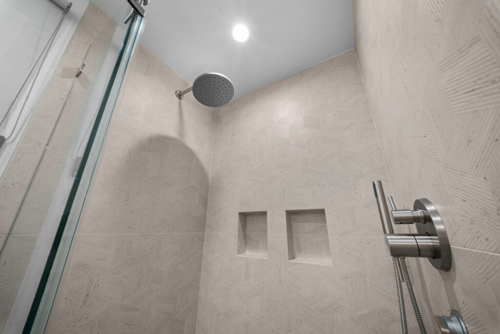 Walk-in shower with textured cream large-format tile, rain showerhead, and dual recessed niches by RnD Builders Inc