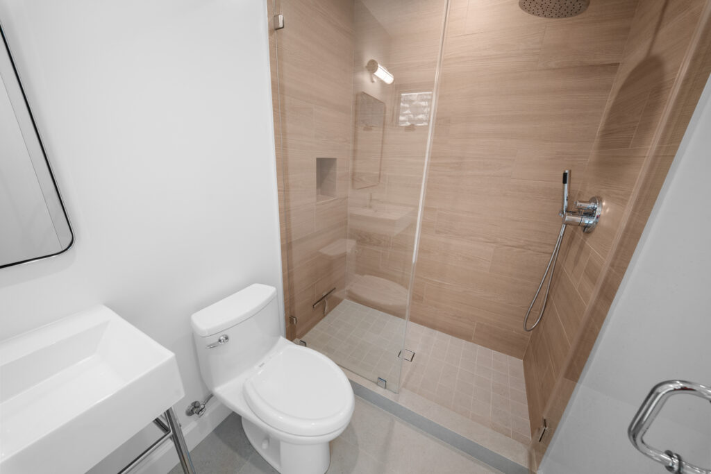 Modern bathroom with frameless glass shower, wood-look tile walls, and white toilet by RnD Builders Inc
