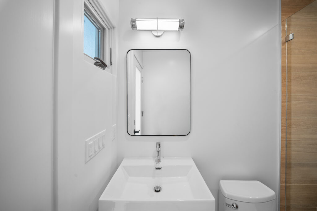 Modern white bathroom with wall-mount sink, black-framed mirror, and wood-look tile shower by RnD Builders Inc