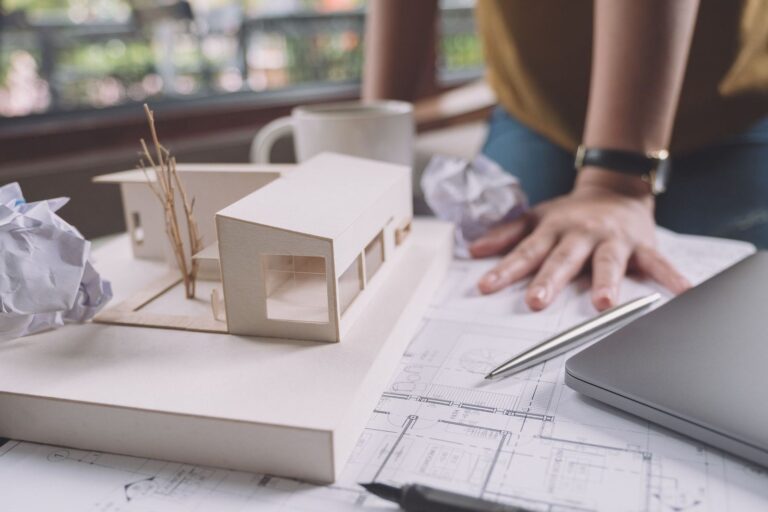 Architectural model of a house, blueprints, and a laptop, on a desk. Architectural plans by RnD Builders