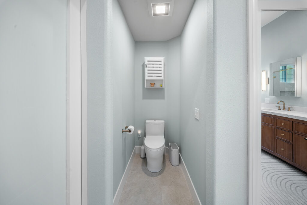 Narrow water closet with white one-piece toilet, louvered wall cabinet, and light blue textured walls by RnD Builders Inc