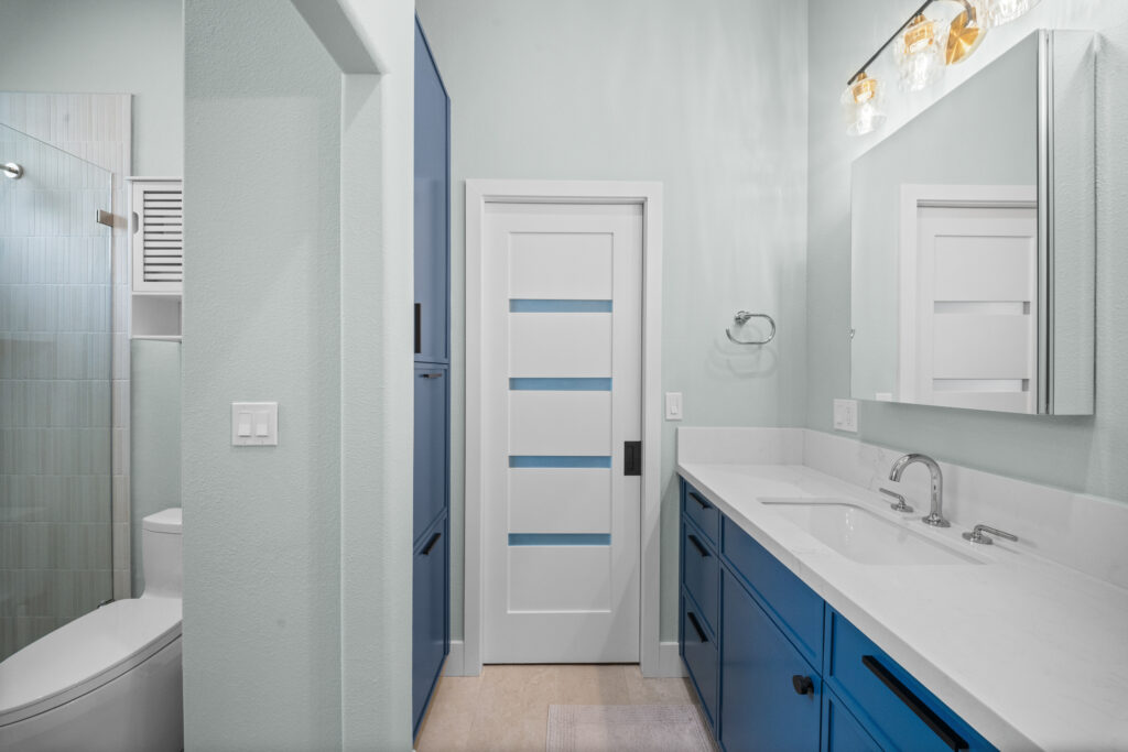 Modern bathroom with blue vanity cabinets, white quartz countertop, and glass-panel door by RnD Builders Inc