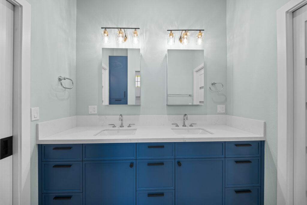 Double-sink bathroom vanity with blue shaker cabinets, white quartz countertop, and gold vanity lights by RnD Builders Inc