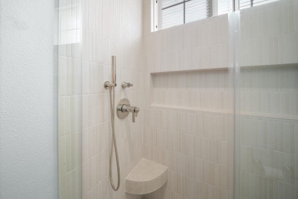 White tile walk-in shower with chrome handheld fixture, built-in niche, and corner shelf by RnD Builders Inc