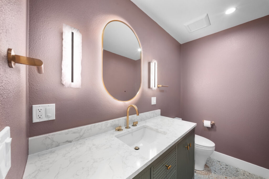 Powder bathroom with mauve walls, marble vanity top, brass fixtures, and oval backlit mirror by RnD Builders Inc