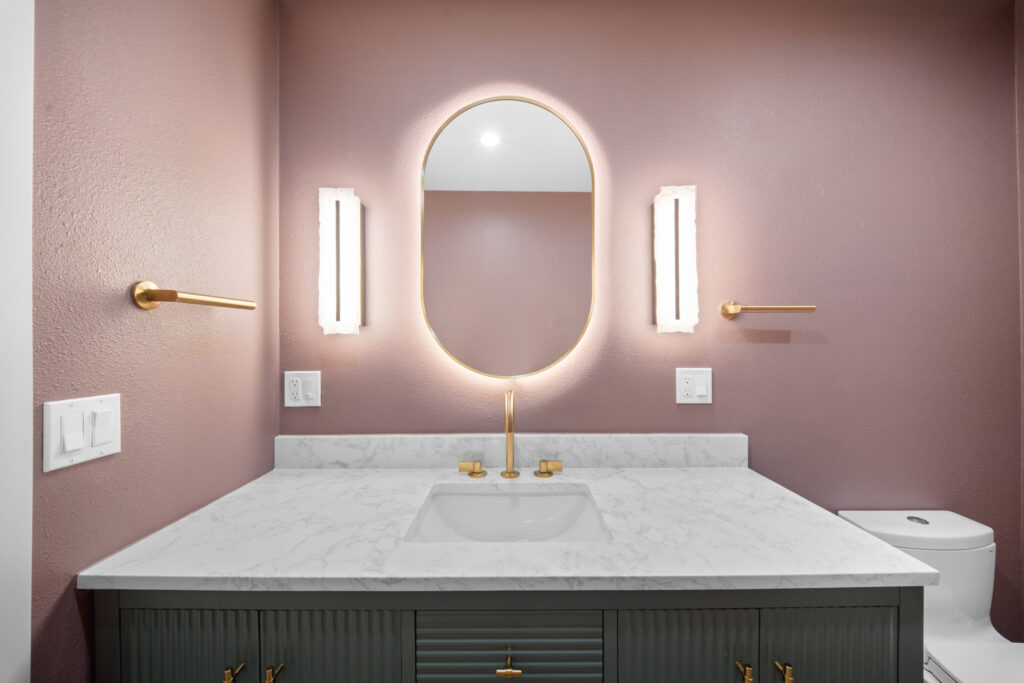 Mauve bathroom vanity with dark green fluted cabinet, marble countertop, and backlit oval mirror by RnD Builders Inc