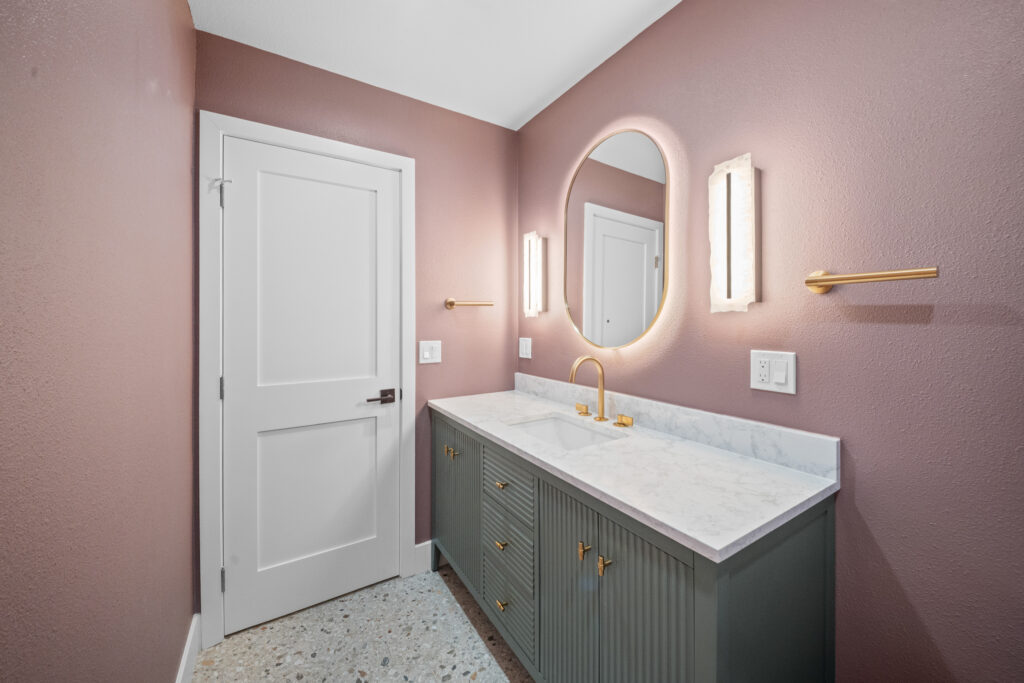 Bathroom with sage green fluted vanity, marble countertop, gold fixtures, and backlit oval mirror by RnD Builders Inc