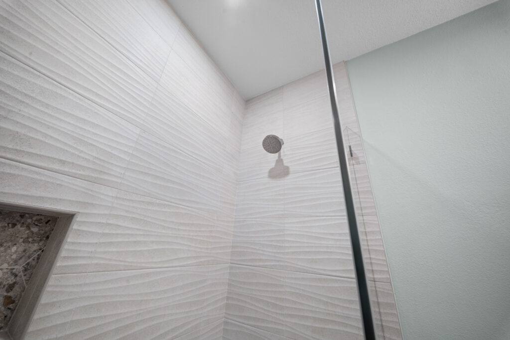 Walk-in shower with white wave-textured wall tiles, round showerhead, and frameless glass panel by RnD Builders Inc