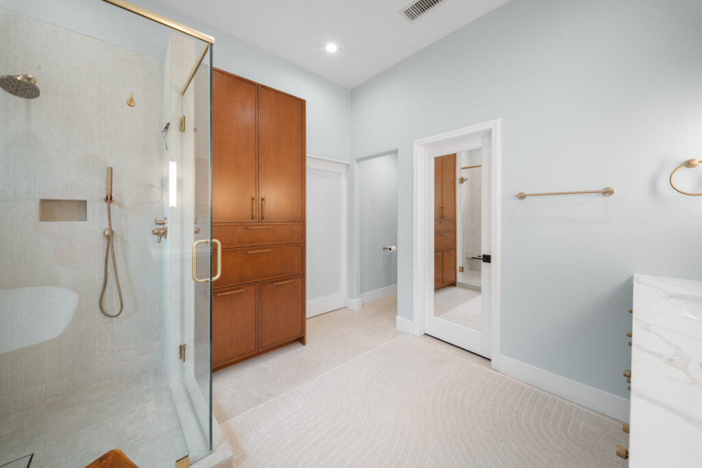 Luxury primary bathroom with glass shower enclosure, warm wood cabinetry, and brushed gold fixtures by RnD Builders Inc