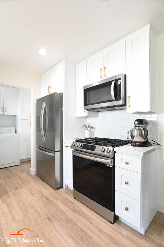 White shaker kitchen with stainless steel appliances, gold hardware, and light wood flooring by RnD Builders Inc