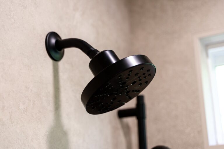 Close-up of matte black rain showerhead mounted on beige stone-look tile wall by RnD Builders Inc