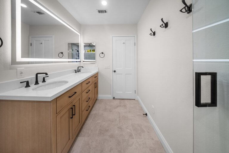 Double-sink master bathroom with warm wood vanity, white quartz countertop, and backlit mirrors by RnD Builders Inc