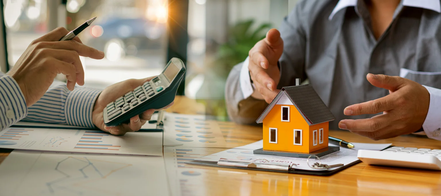 Two people at a desk with a miniature house model, calculator, and financial documents by RnD Builders Inc