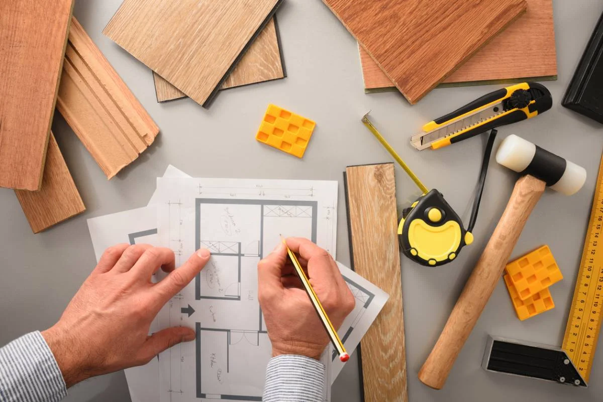 Contractor reviewing floor plan blueprints surrounded by wood flooring samples and construction tools by RnD Builders Inc