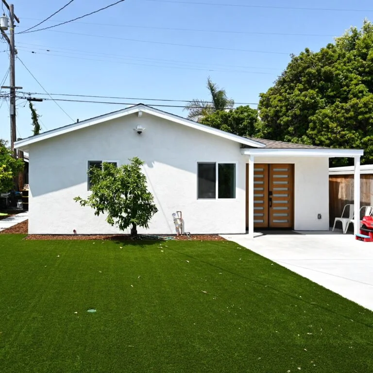 Single-story white stucco home with modern wood double entry door and artificial turf front yard by RnD Builders Inc