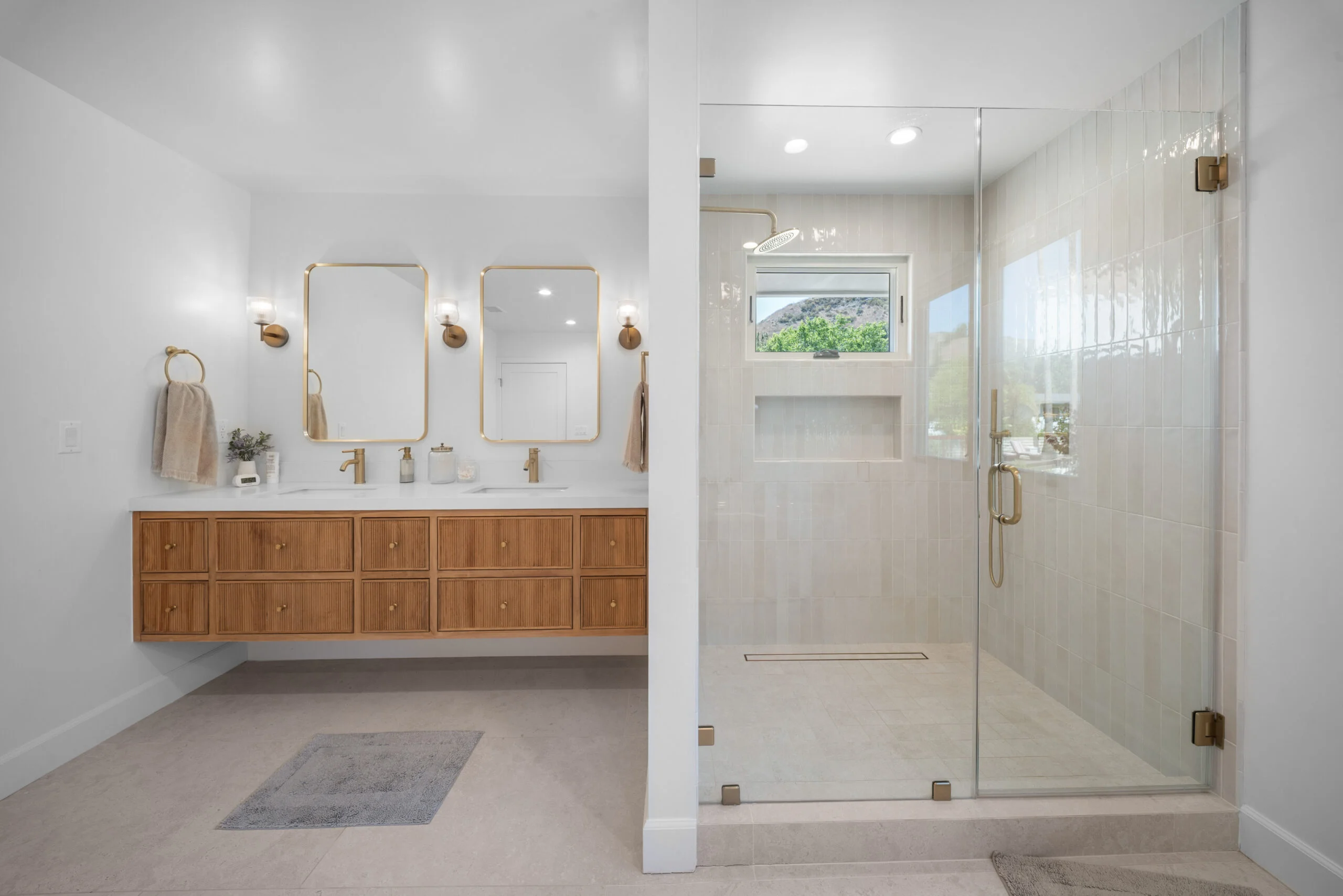 Bright primary bathroom with floating fluted wood vanity, dual gold mirrors, and frameless glass shower by RnD Builders Inc
