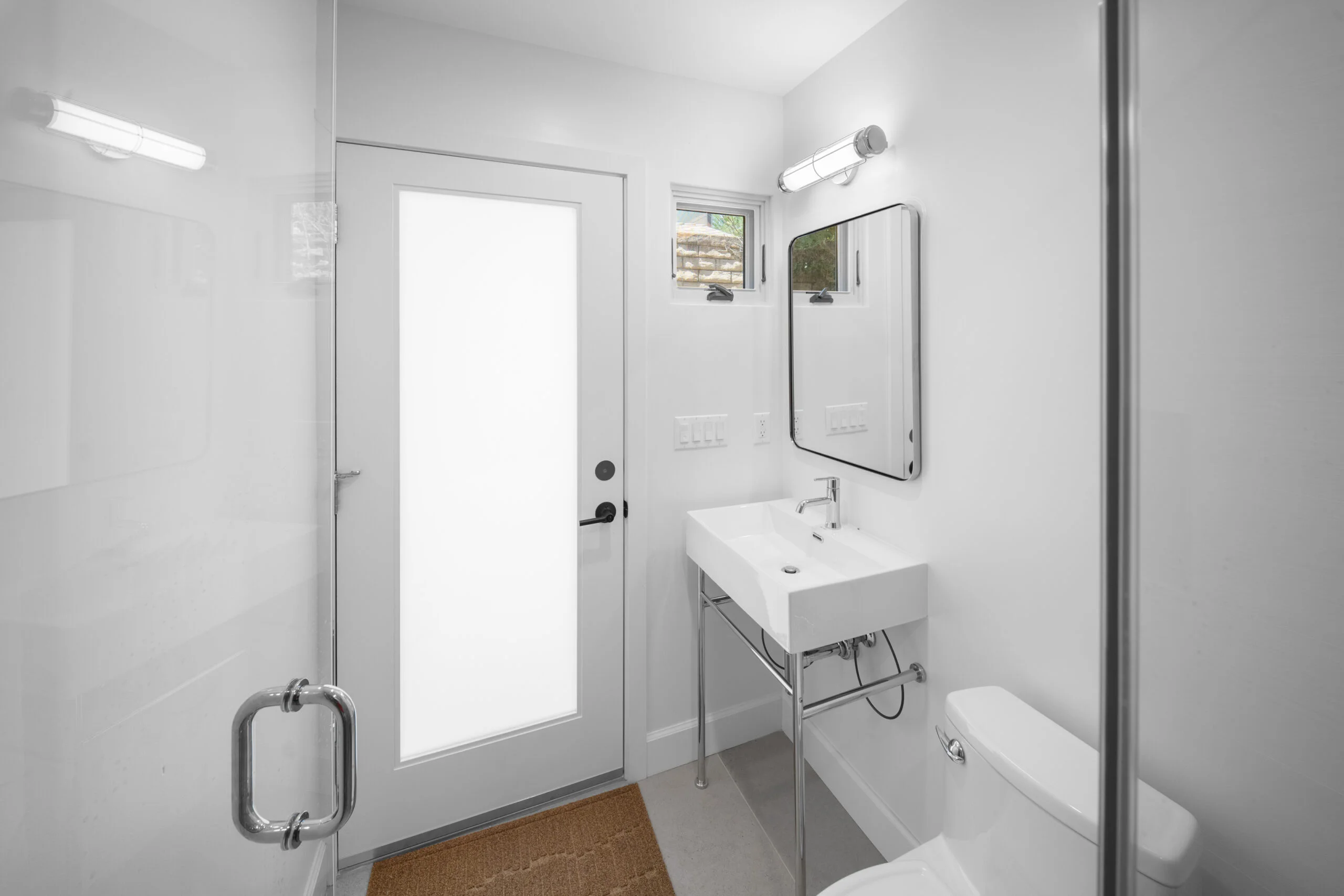 Modern white bathroom with console sink, glass shower door, and frosted glass entry door by RnD Builders Inc