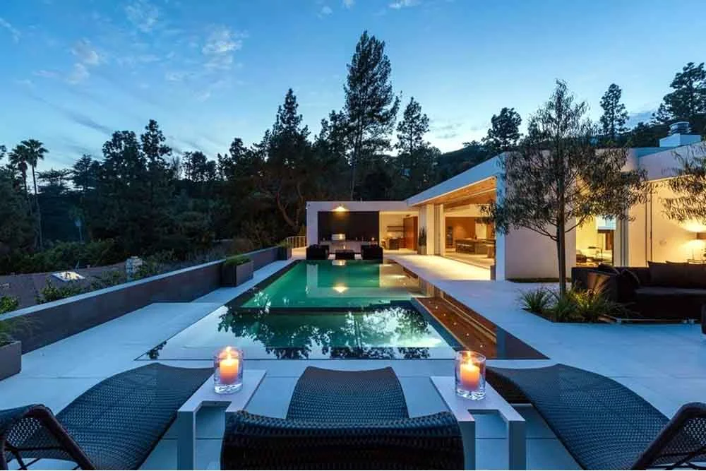 Luxury modern home with rectangular pool, candle lit lounge chairs, and open indoor-outdoor living at dusk by RnD Builders Inc