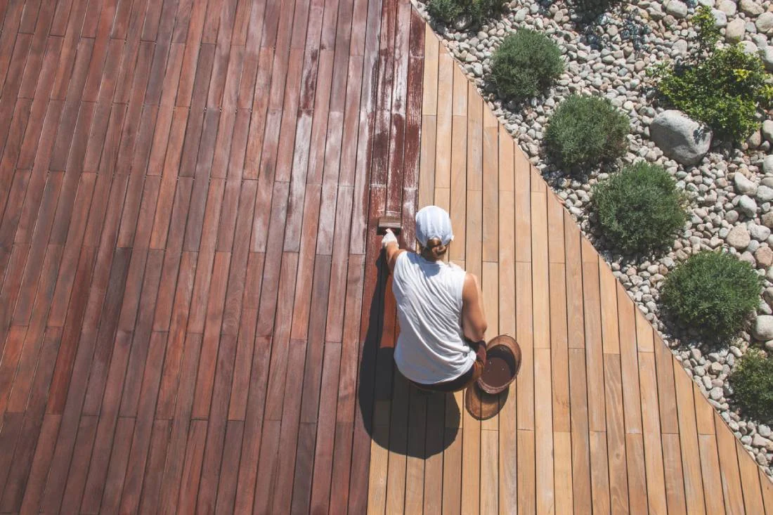 Worker applying wood stain to a large hardwood deck beside a gravel and shrub landscape by RnD Builders Inc