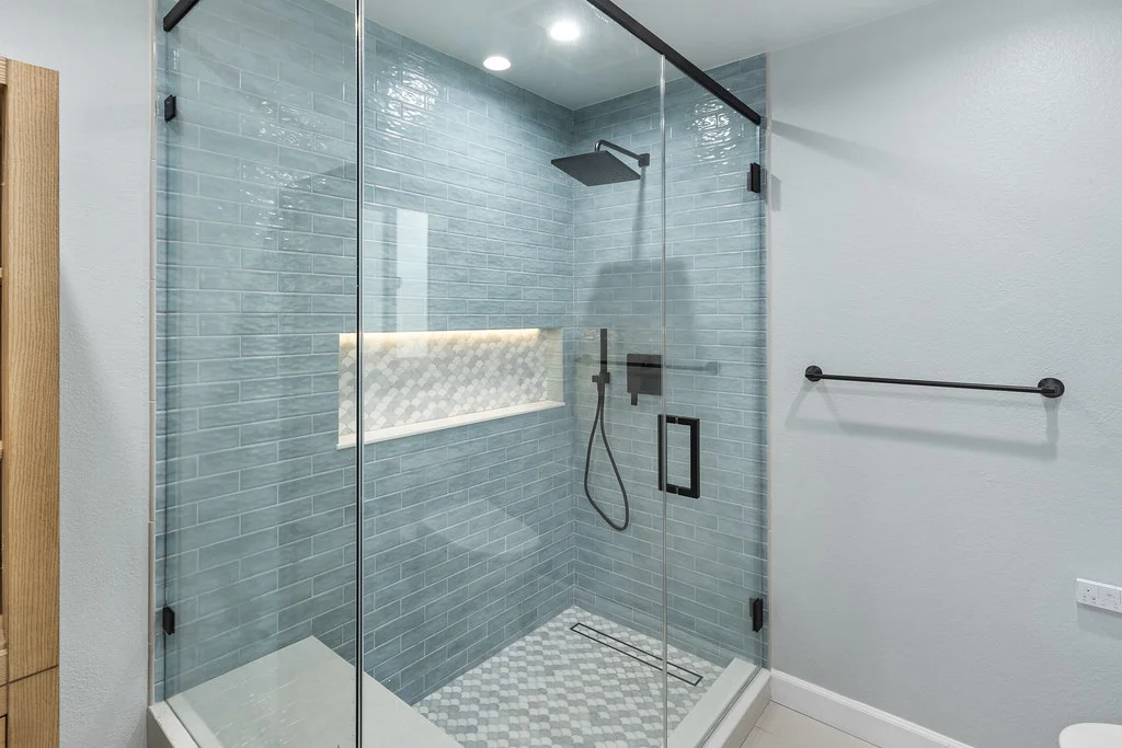 Walk-in glass shower with blue subway tile, matte black fixtures, and illuminated niche by RnD Builders Inc