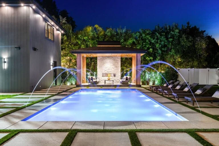 Illuminated rectangular pool with arc water jets, covered pavilion, and stone fireplace at night by RnD Builders Inc