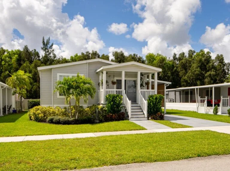 Light gray manufactured home with white covered front porch, steps, and tropical landscaping by RnD Builders Inc