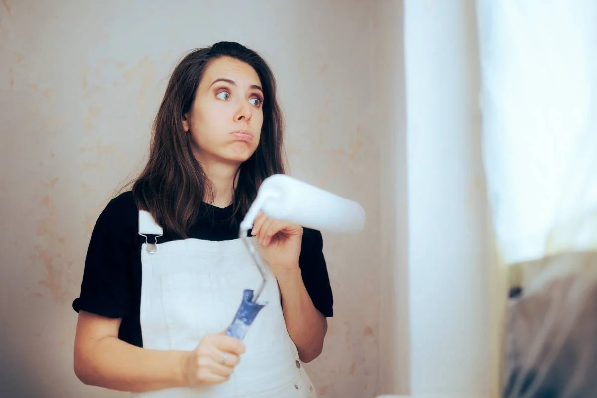 Woman in white overalls holding a paint roller near an unpainted interior wall by RnD Builders Inc